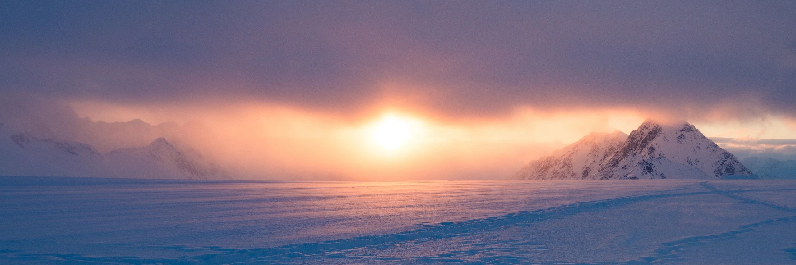 Snow-covered mountain landscape at sunrise, featuring soft pastel colors in the sky and a visible trail in the snow, reflecting the adventure and beauty of peak exploration for climbers and hikers.