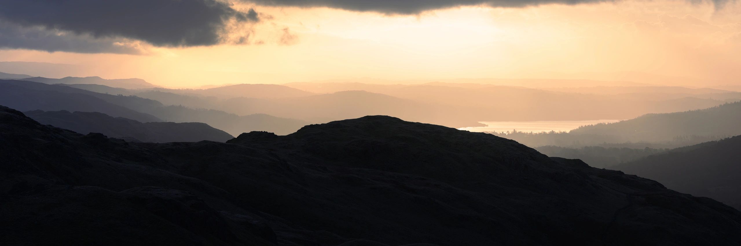 Mountain landscape at sunset, showcasing layered hills and valleys, with soft light illuminating the distant peaks and a tranquil body of water in the background, reflecting the essence of adventure and exploration for climbers and hikers.