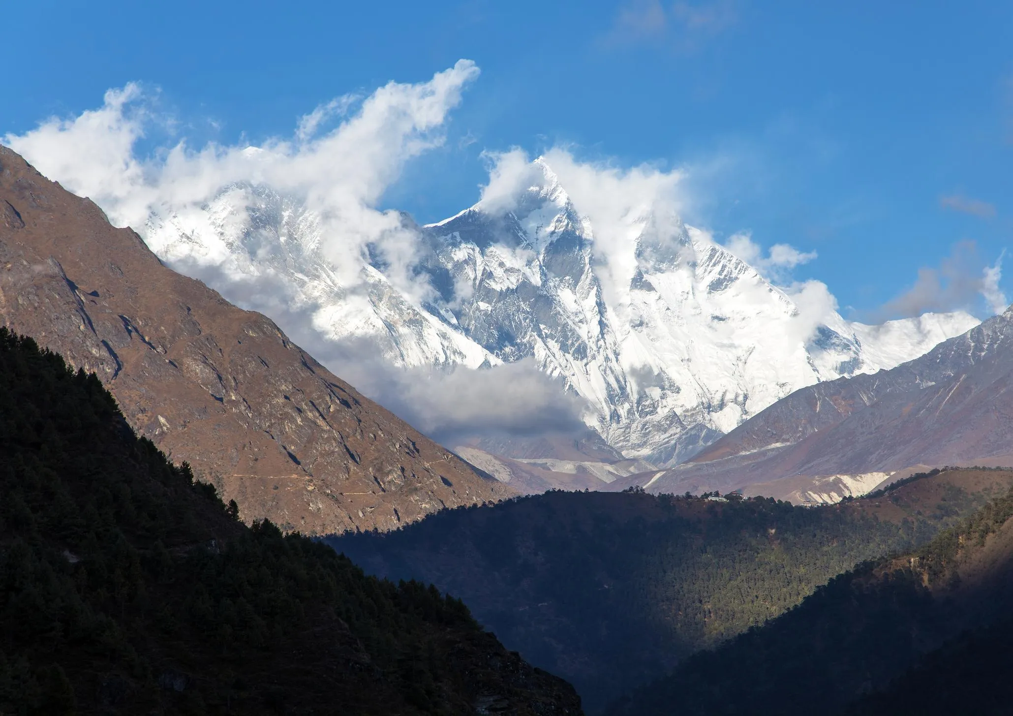 Snow-capped peaks of the Himalayas under a blue sky, showcasing the majestic Lhotse mountain range, surrounded by rugged terrain and clouds.