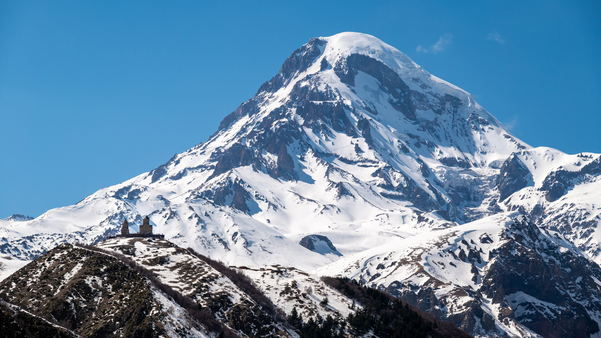 Mount Kazbek summit at 5054 meters in the Greater Caucasus showing the glaciated dormant stratovolcano above Stepantsminda Georgia with the iconic Gergeti Trinity Church visible in the foreground during 2026 climbing season