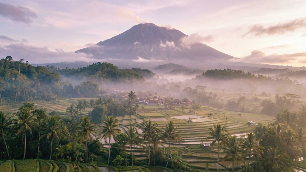 Mount Agung volcano rising above lush green rice terraces and palm trees in Bali, Indonesia, during a misty sunrise.