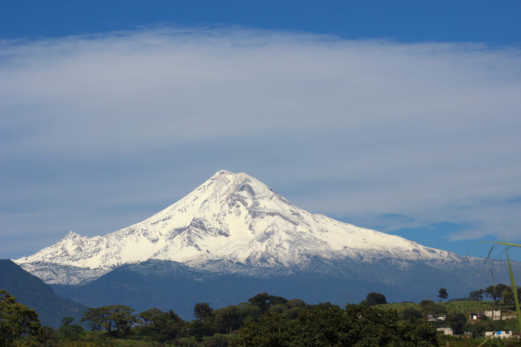 Snow-capped Pico de Orizaba (Citlaltépetl) towering against a clear blue sky, surrounded by green hills and vegetation, representing Mexico's highest mountain and popular climbing destination.