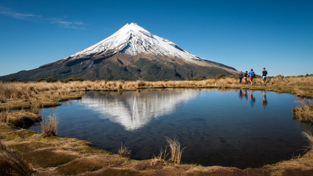 Mount Taranaki reflected in a calm pond, with hikers walking along the grassy foreground in Egmont National Park, New Zealand.