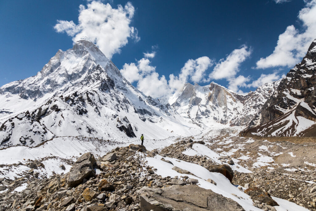 Snow-covered Mount Meru towering over rocky terrain with a lone trekker in the foreground, showcasing the challenging landscape of the Indian Himalayas.