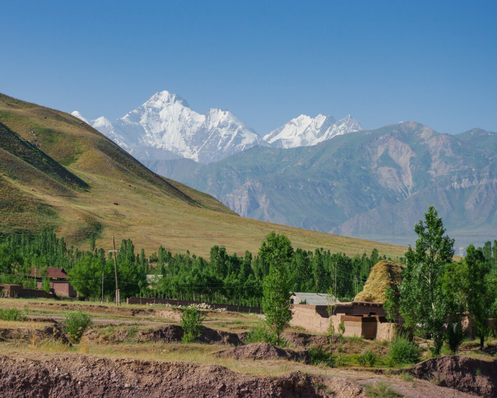 Ismoil Somoni Peak in the background, towering snow-capped mountains, lush green valley with trees and rustic buildings in the foreground, highlighting Tajikistan's natural beauty and mountaineering landscape.