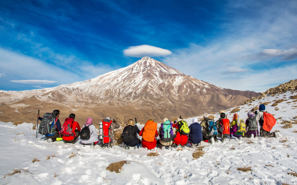 Group of climbers in colorful gear seated on snowy terrain, overlooking Mount Damavand, the highest peak in Iran, under a blue sky with clouds.