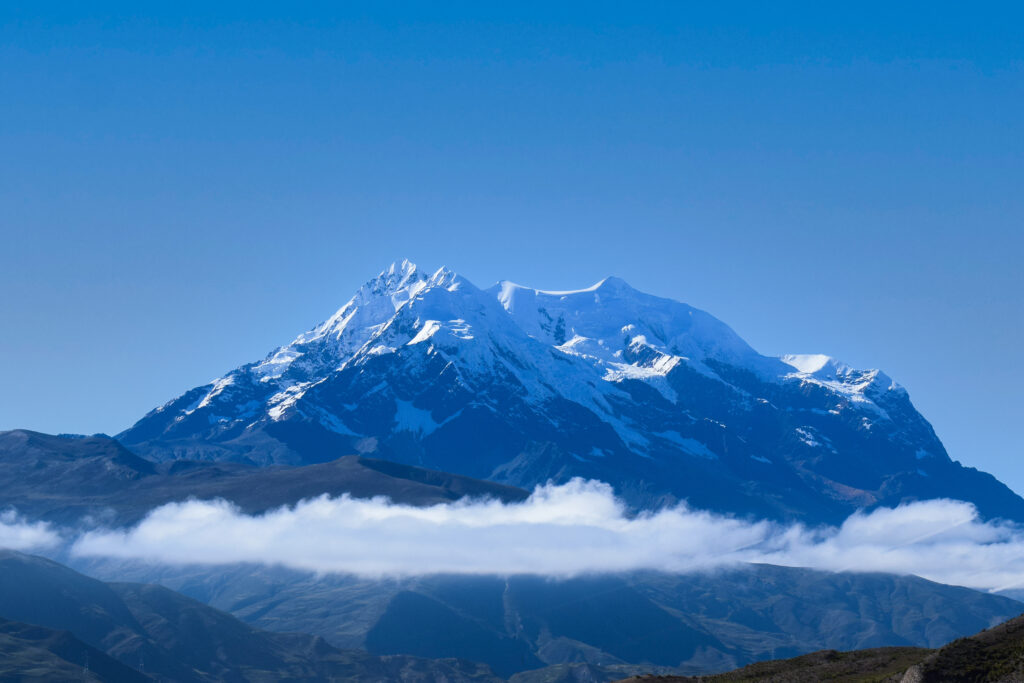 Illimani mountain in Bolivia, showcasing snow-capped peaks against a clear blue sky, with clouds hovering over the lower slopes, emphasizing its prominence as a high-altitude climbing destination.