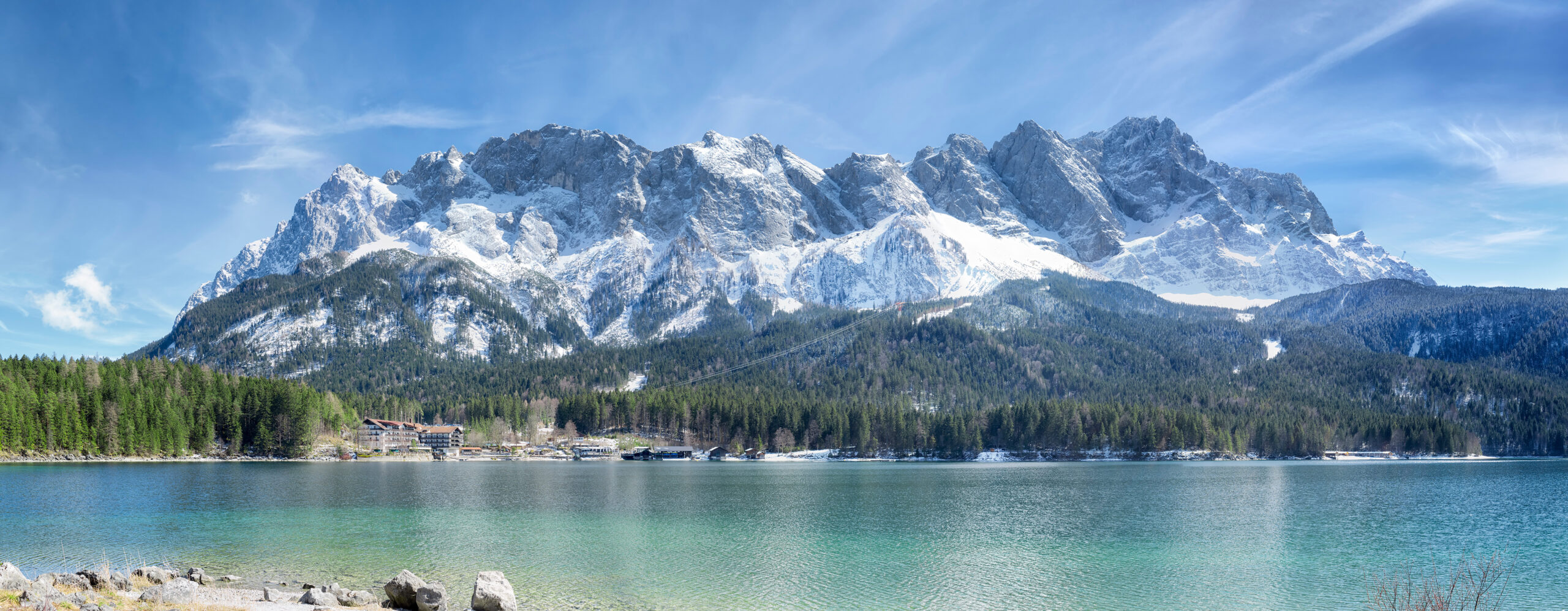 Zugspitze mountain range in Bavaria, Germany, featuring snow-capped peaks, lush green forests, and a serene lake in the foreground.