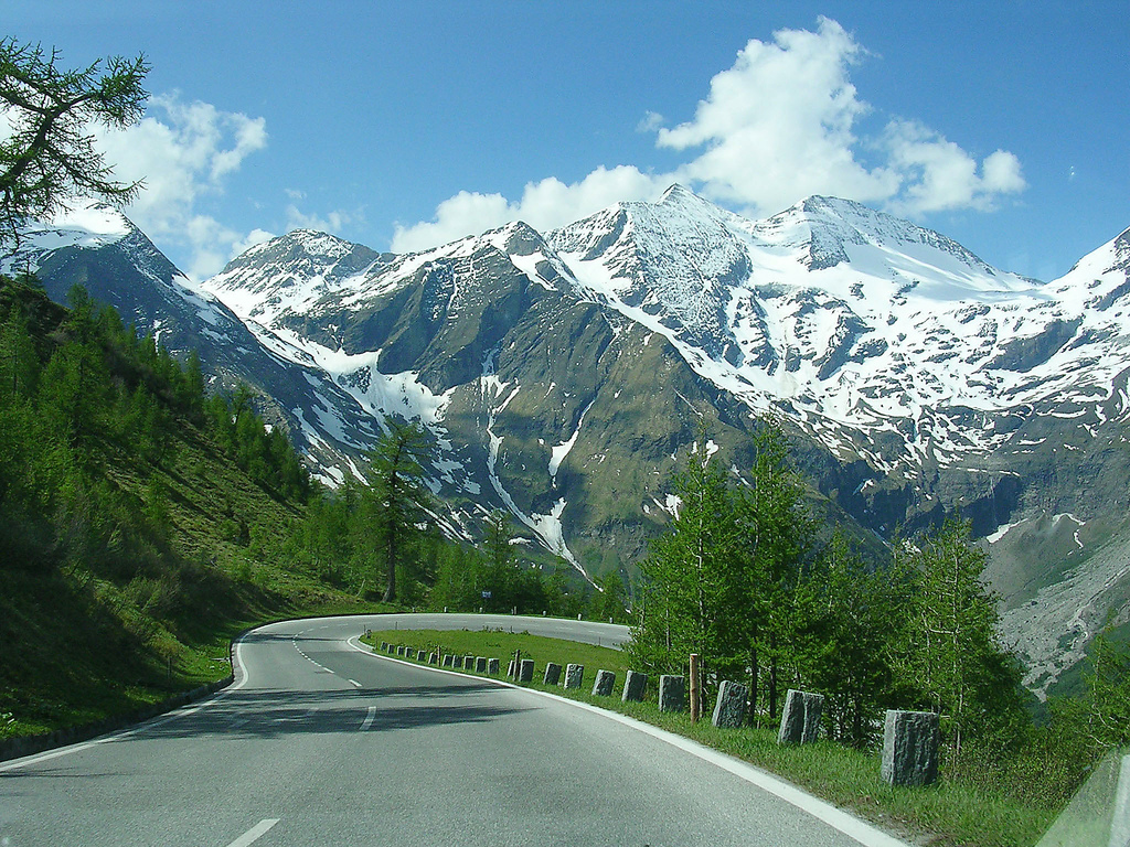 Scenic view of Grossglockner mountain in Austria, featuring winding road, lush greenery, and snow-capped peaks under a blue sky, ideal for climbers and outdoor enthusiasts.