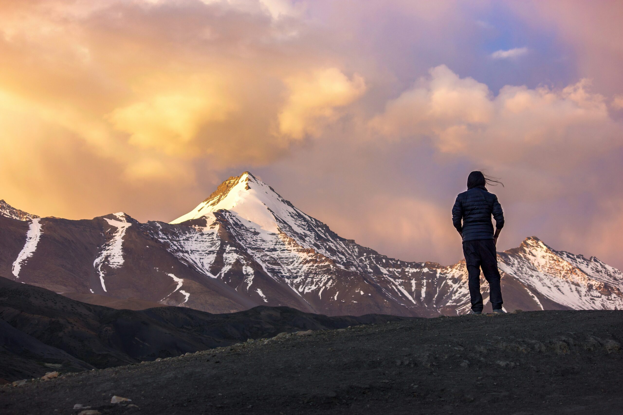 Person standing on rocky terrain, overlooking snow-capped mountains at sunset, illustrating the beauty and challenges of trekking and mountaineering.