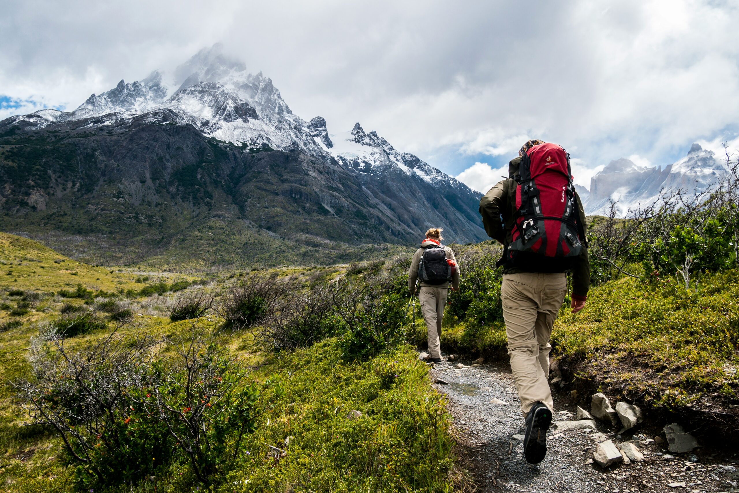 Hikers trekking on a mountain path with backpacks, surrounded by lush greenery and snow-capped peaks, illustrating outdoor adventure and exploration.