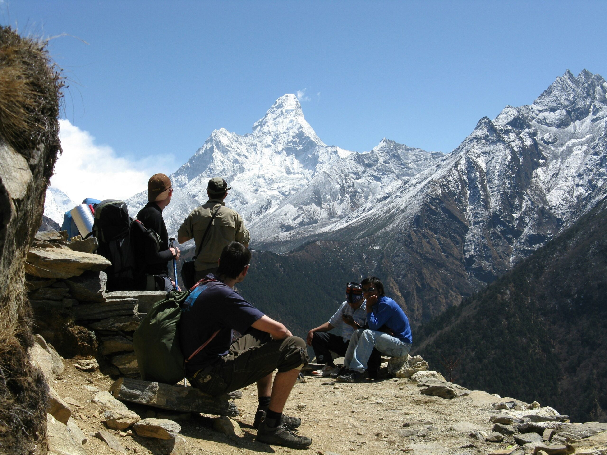 Climbers resting on rocky outcrop with backpacks, overlooking snow-capped Ama Dablam mountain in Nepal, highlighting the alpine environment and trekking conditions.