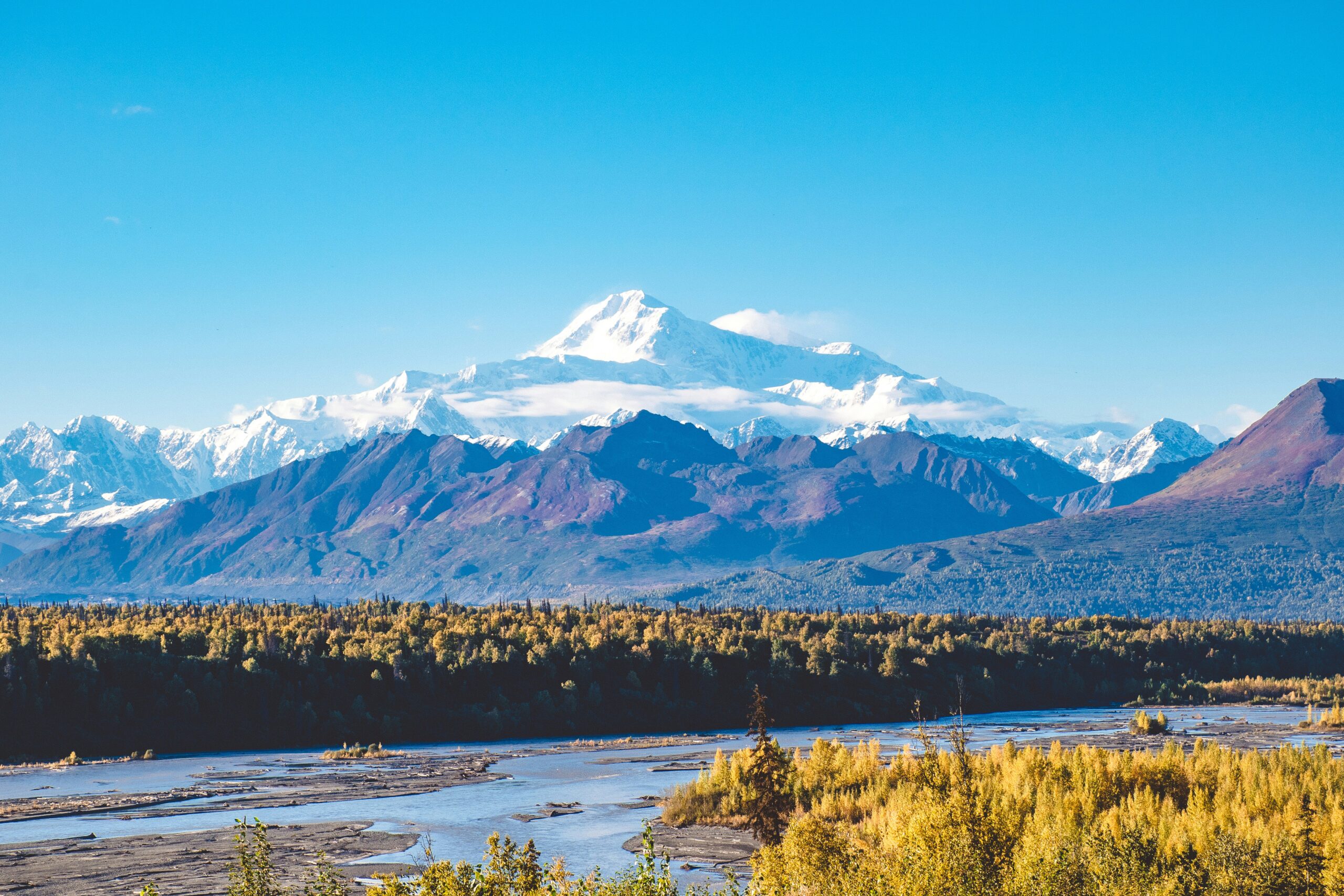 Majestic view of snow-capped peak in the distance, surrounded by rugged mountains and vibrant autumn foliage, representing the breathtaking landscape of climbing expeditions.