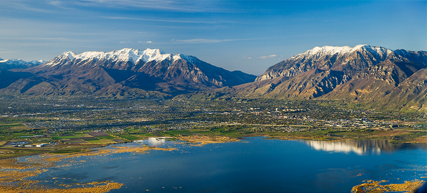 Aerial view of Utah County's Wasatch Range, featuring snow-capped peaks, lush valleys, and a reflective lake, highlighting prime climbing locations for summer adventures.