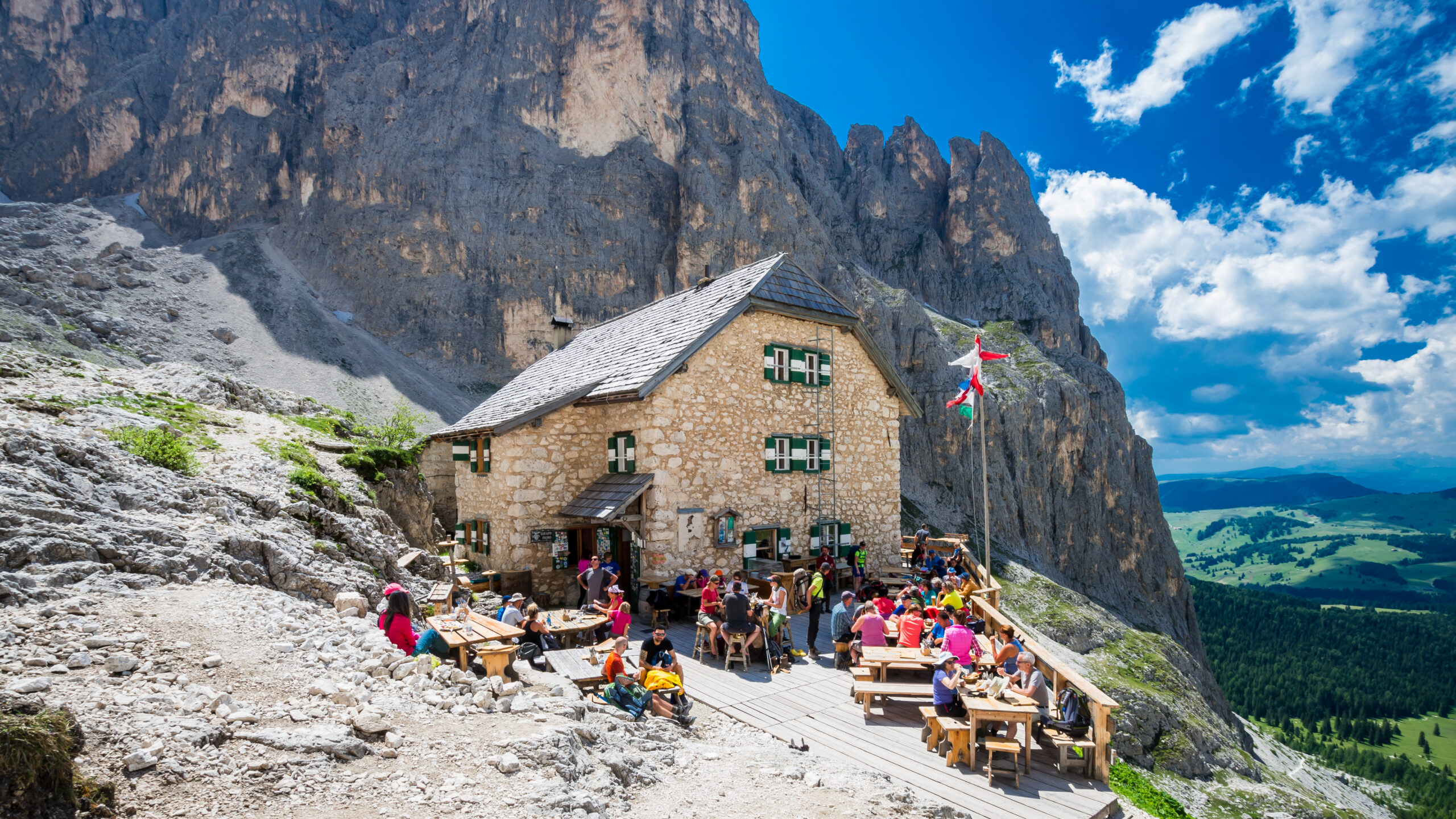 A mountain refuge with hikers resting on a terrace below high peaks